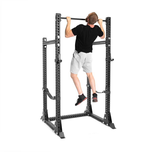 A man wearing a black shirt and gray shorts does a pull-up on the Bells of Steel Manticore Half Rack - Prebuilt (3" x 3", 1" Holes), set against a white background—perfect for any home gym.