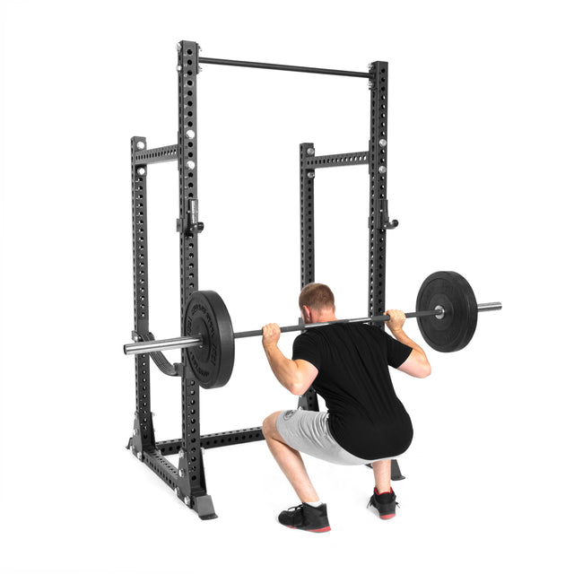 A man in a black shirt and gray shorts performs a barbell squat inside the Bells of Steel Manticore Half Rack - Prebuilt (3" x 3", 1" Holes) set against a white background—an ideal addition to any home gym.