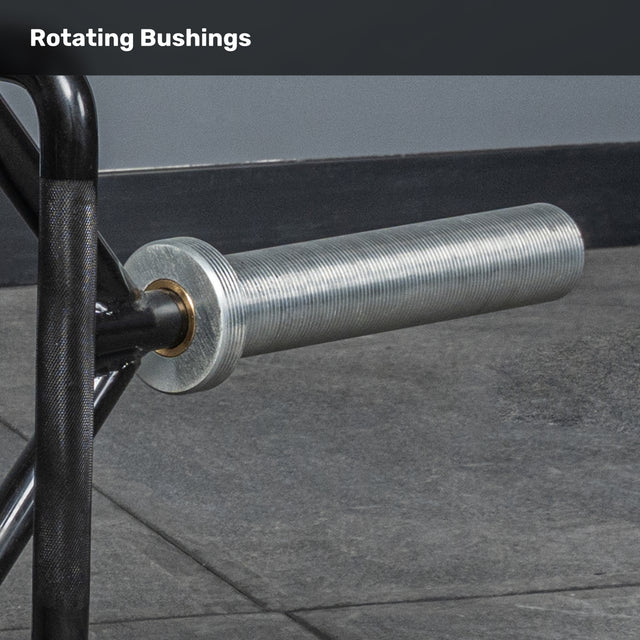 The image shows a close-up of the Bells of Steel Open Trap Bar's sleeve with rotating bushings on its textured metal bar, resting on a gym floor. “Rotating Bushings” is displayed at the top.