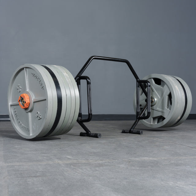 The Bells of Steel Open Trap Bar / Hex Bar, loaded with heavy plates, sits on a gray gym floor by a plain wall—this essential strength-training tool is ready for your next lift.
