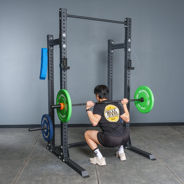 A person using the Bells of Steel Hydra Half Rack - Prebuilt (3" x 3", ⅝" Holes) performs a barbell squat with colored plates, showing how this modular rack can elevate any home gym space.