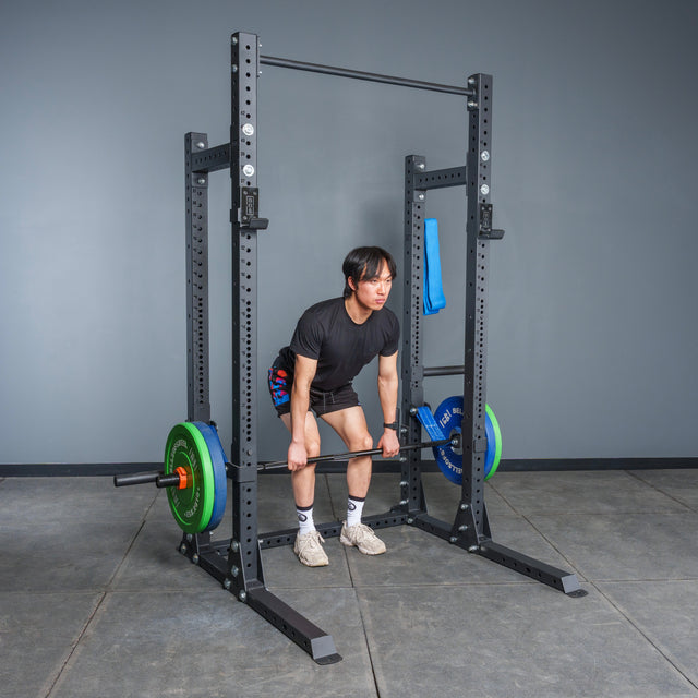 Wearing athletic gear, a person deadlifts a loaded barbell inside a Bells of Steel Hydra Half Rack - Prebuilt (3" x 3", ⅝" Holes) in a home gym with gray walls, modular features, and a concrete floor.