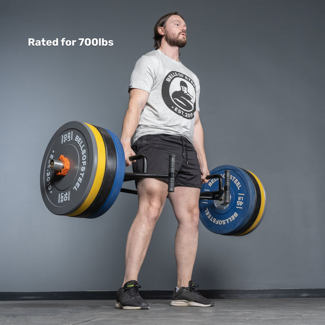 A man lifts the Bells of Steel Open Trap Bar / Hex Bar with large weight plates in a gym. He wears a grey t-shirt, black shorts, and black sneakers. Text on image: "Rated for 700lbs.
