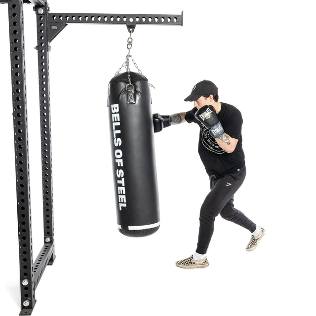 Wearing boxing gloves, a black shirt, black pants, and a cap, a person punches a Bells of Steel USA Heavy Bag Set in a white studio—a dynamic scene for any combat sports fan.