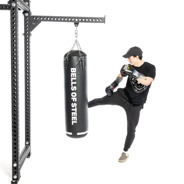A person wearing black clothes and boxing gloves kicks a Heavy Bag Set by Bells of Steel USA, which hangs from a power rack against a white background.