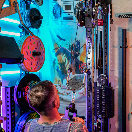 A person with short hair uses a home gym cable machine in a vibrant space featuring weight plates, a wall poster, boombox, neon lights, and the Bells of Steel USA Kaizen Multi-Link for versatile workouts.