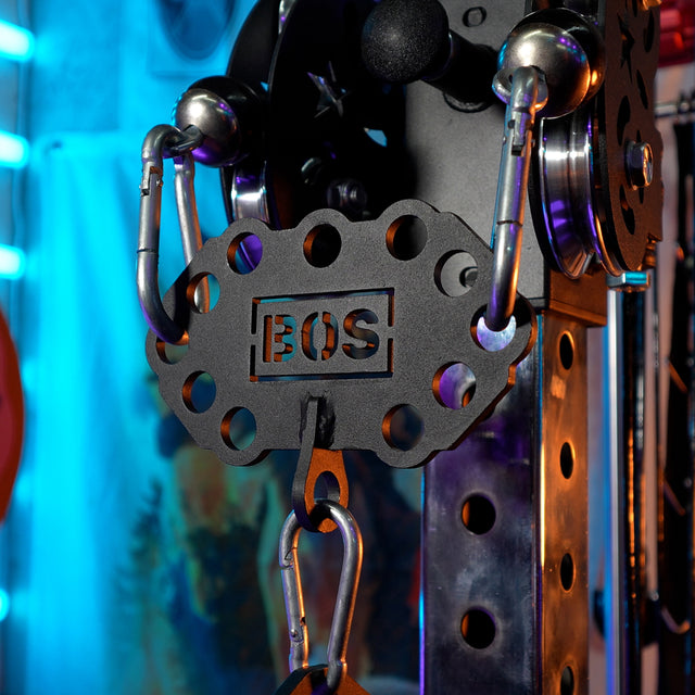 Close-up of a black metal gym attachment labeled "BOS" connected to a cable machine using the Kaizen Multi-Link from Bells of Steel USA, set against neon blue and purple lighting.