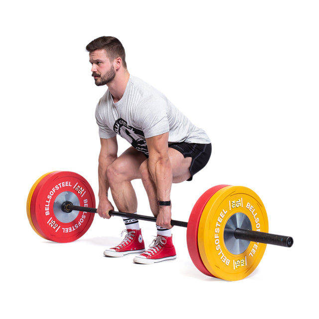 A man in a gray T-shirt, black shorts, and red sneakers prepares to deadlift a barbell loaded with Bells of Steel Boltless KG Competition Bumper Plates, standing against a white background.
