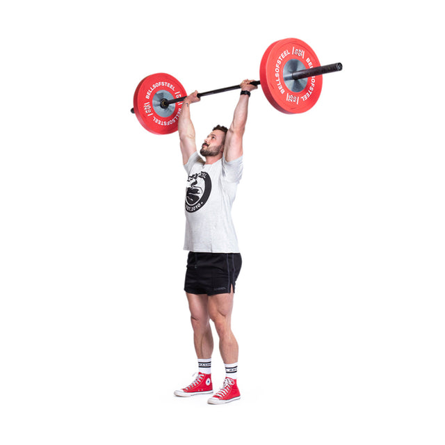 A man in a gray t-shirt, black shorts, and red shoes lifts a barbell loaded with Bells of Steel Boltless KG Competition Bumper Plates overhead, standing tall with arms extended as he performs an Olympic weightlifting move.