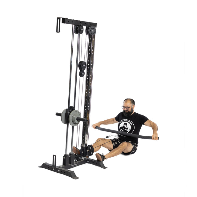A man with glasses and a beard uses the Bells of Steel Fat Bar - Lat Pulldown Bar, pulling it with both hands to build grip strength. He sits on the floor in a black shirt and shorts against a white background.