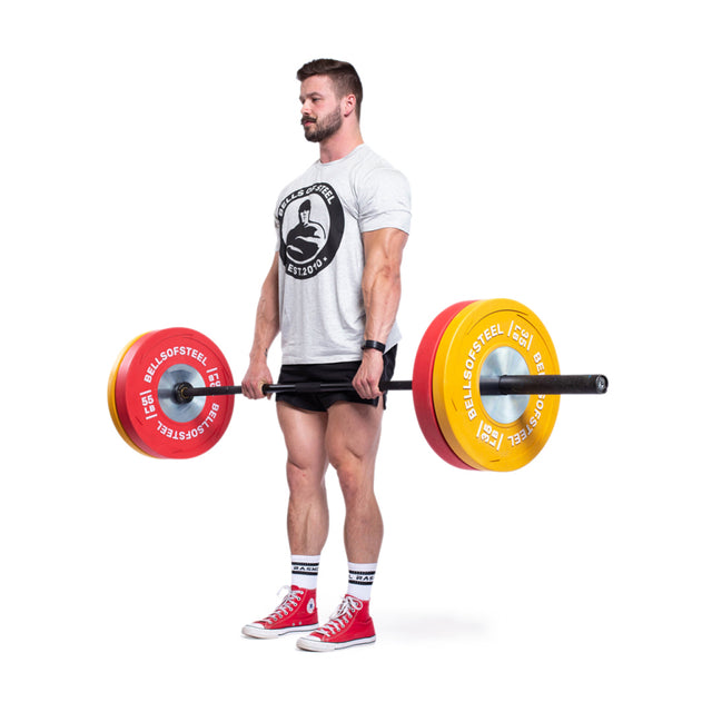 A man lifts a barbell loaded with Bells of Steel Boltless LB Competition Bumper Plates, built to IWF specifications, while wearing a gray t-shirt, black shorts, and red shoes against a white background.
