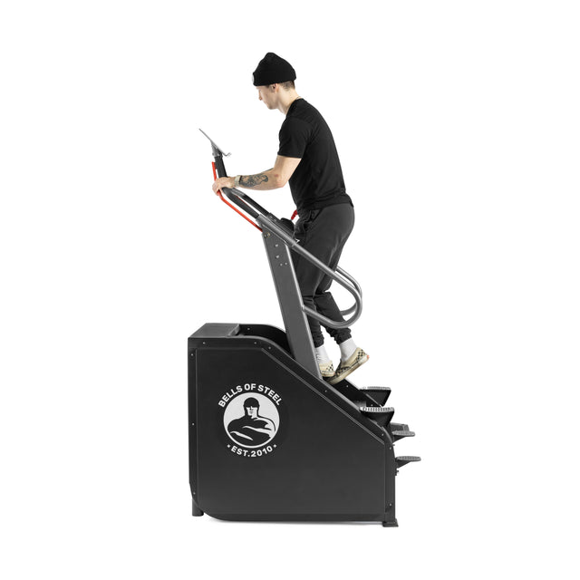 Wearing workout clothes and a beanie, someone uses the black Bells of Steel Sisyphean Stepper - Manual Stair Climber, stepping upward while gripping the handrails—an ideal addition to any home gym cardio lineup.