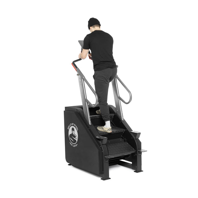 A person in black attire and a beanie uses the Bells of Steel Sisyphean Stepper - Manual Stair Climber with handrails, viewed from behind on a white background—ideal inspiration for your home gym cardio machine setup.