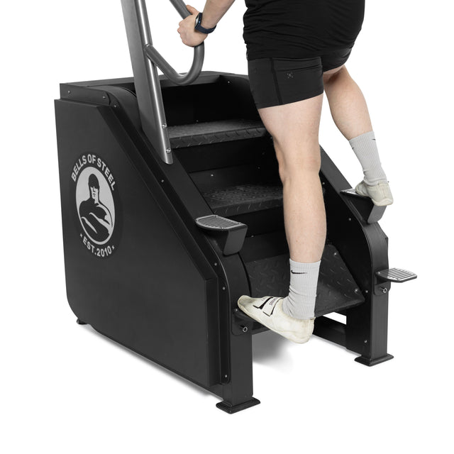 Wearing black shorts and gray socks, a person grips the handrails while using the Bells of Steel Sisyphean Stepper manual stair climber, stepping up on the cardio machine’s stairs.