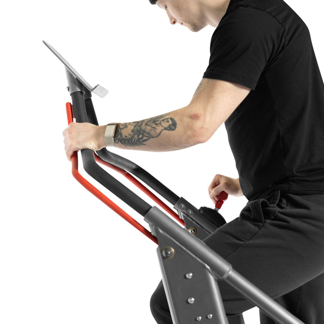 Wearing black workout clothes and a smartwatch, a tattooed user grips the red and black handles of the Bells of Steel Sisyphean Stepper – Manual Stair Climber, exercising on this cardio machine against a white background.