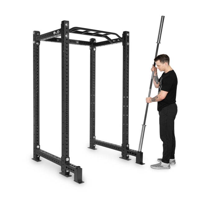 A man in black athletic clothing holds a barbell beside a Bells of Steel power rack with Front Foot Stabilizers, barbell storage, multiple attachment holes, and a multi-grip pull-up bar.