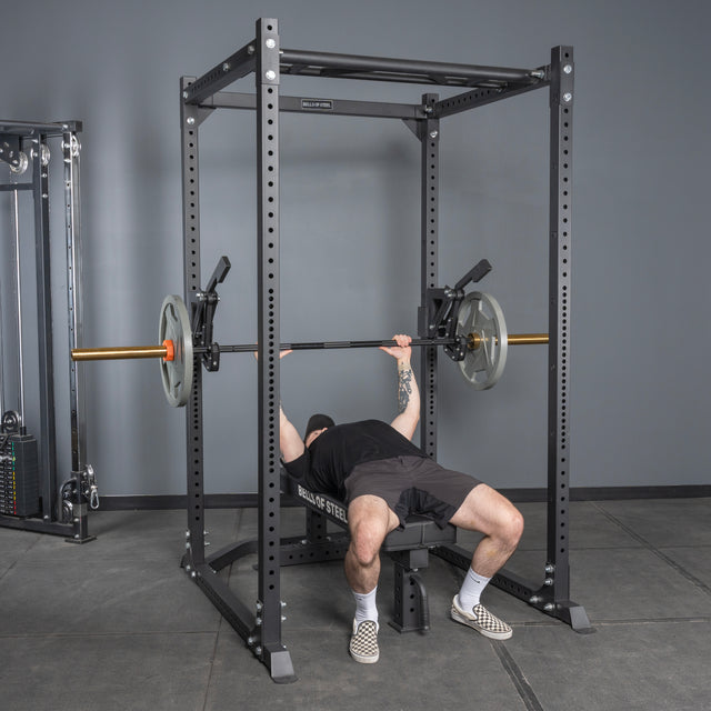 A person wearing black shorts and checkered shoes bench presses a barbell in a black power rack with the Bells of Steel Monolift Rack Attachment—a space-saving solution for efficient workouts.