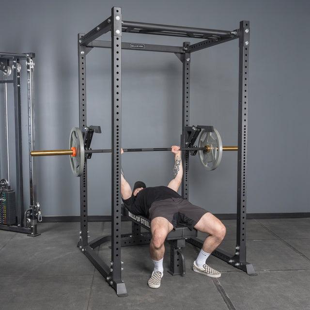 Wearing black shorts and a T-shirt, a person bench presses with a barbell inside a power rack featuring the Bells of Steel Monolift Rack Attachment, creating a more space-saving gym setup.