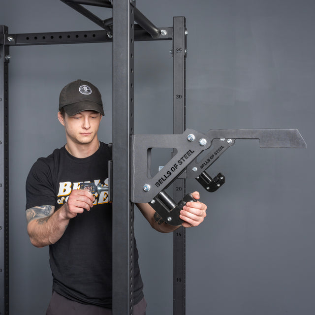 A person wearing a black Bells of Steel shirt and cap installs a Bells of Steel Monolift Rack Attachment onto a squat rack in a gym.