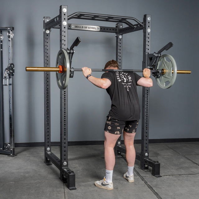 A person prepares for a squat in the gym using the Bells of Steel Monolift Rack Attachment, holding a weighted barbell across their upper back and making final adjustments before lifting.