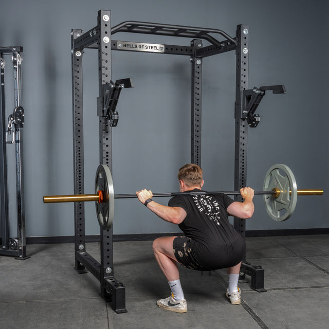 Wearing athletic clothing, a person performs a barbell back squat in a gym power rack equipped with the Bells of Steel Monolift Rack Attachment, using its space-saving design to securely hold the barbell during squats.
