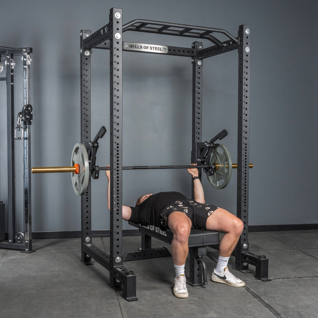 A person is bench pressing with weight plates on a bench inside a power rack equipped with the Bells of Steel Monolift Rack Attachment, in a gym with gray walls and concrete flooring. The attachment provides space-saving bar adjustments.