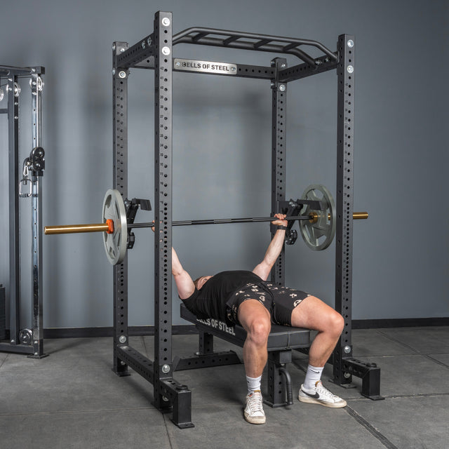 A person uses the Bells of Steel Monolift Rack Attachment for a bench press, easily adjusting the bar inside a power rack for efficient lifts in a space-saving gym setup.