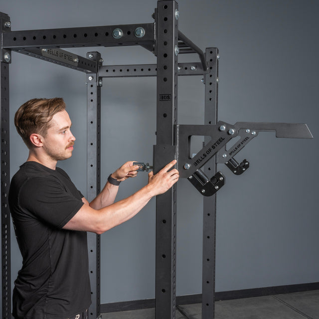 A man in a black t-shirt adjusts a black metal power rack in the gym, focusing on attaching the space-saving Bells of Steel Monolift Rack Attachment against a gray wall.