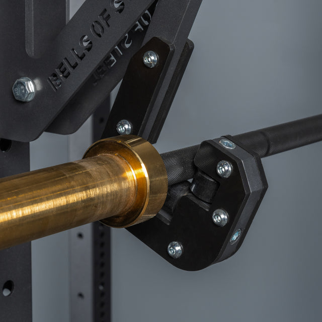 Close-up of a Bells of Steel Monolift Rack Attachment, featuring a brass-colored barbell sleeve and heavy-duty black metal rack with multiple bolts and bar adjustments, designed for space-saving gym setups.