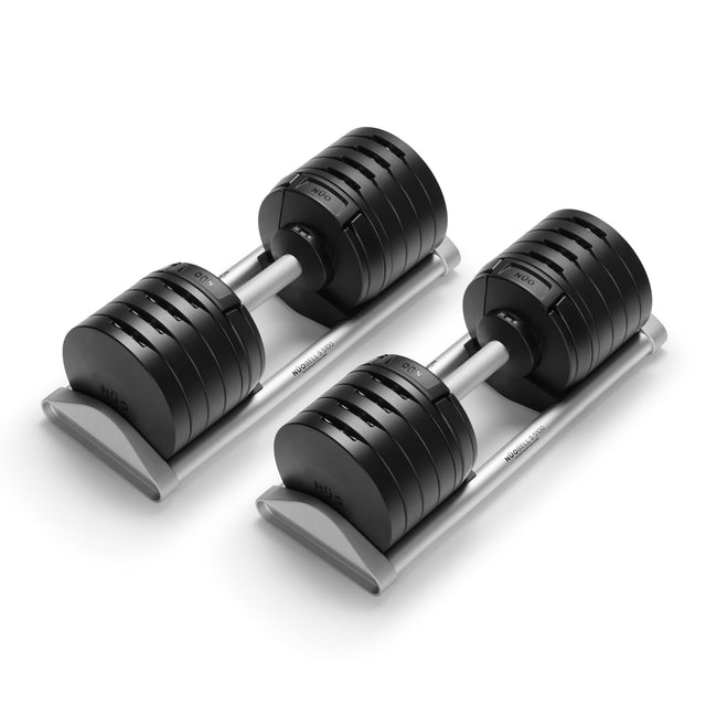 Two Bells of Steel USA NÜOBELL-S Adjustable Dumbbells with black weight plates and metallic handles are displayed on a silver stand. Clearly labeled increments make them ideal for any home gym setup.