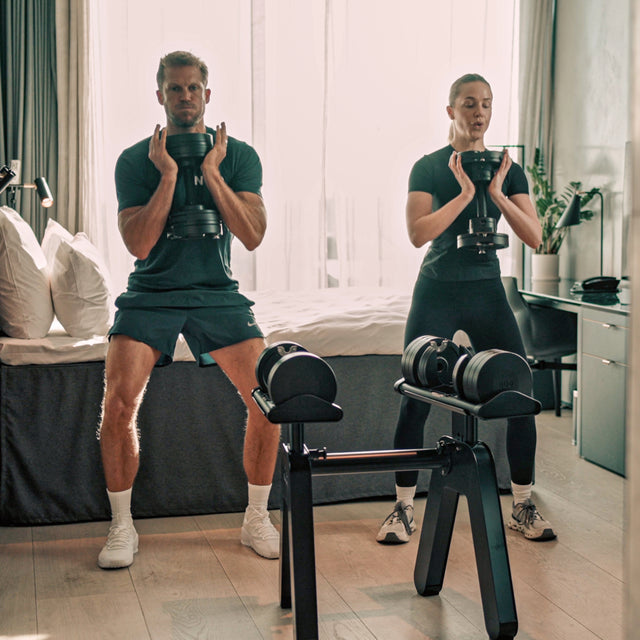 Two people in workout gear do squats in a sunlit bedroom, each holding a Bells of Steel USA NÜOBELL-S Adjustable Dumbbell. A matching rack sits in front of them, creating the perfect home gym atmosphere.