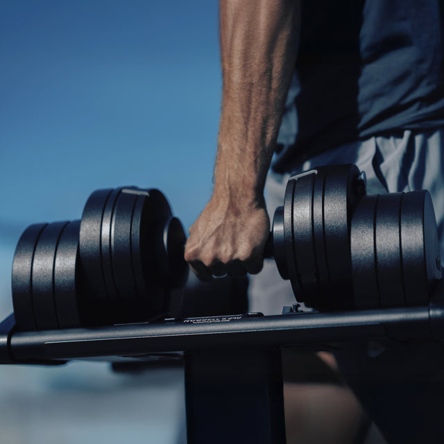 A close-up shows a muscular arm gripping the handle of a NÜOBELL-S Adjustable Dumbbell by Bells of Steel USA on a rack in a home gym, with a blurred outdoor view and blue sky in the background.