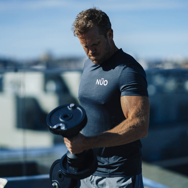 A man lifts a NÜOBELL-S Adjustable Dumbbell by Bells of Steel USA outdoors on a sunny day, with buildings and a blue sky blurred in the background.
