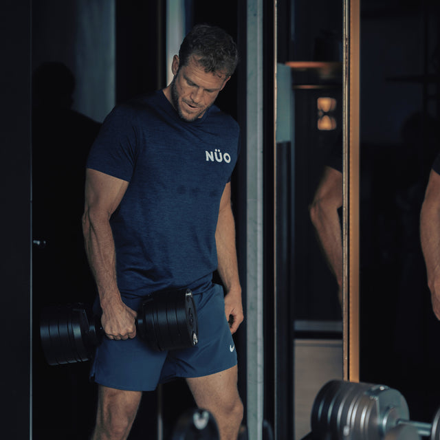 A man in a navy blue NUO shirt and shorts lifts the NÜOBELL-S Adjustable Dumbbells by Bells of Steel USA in a dimly lit gym, focusing downward as a large mirror reflects his image and surrounding equipment.