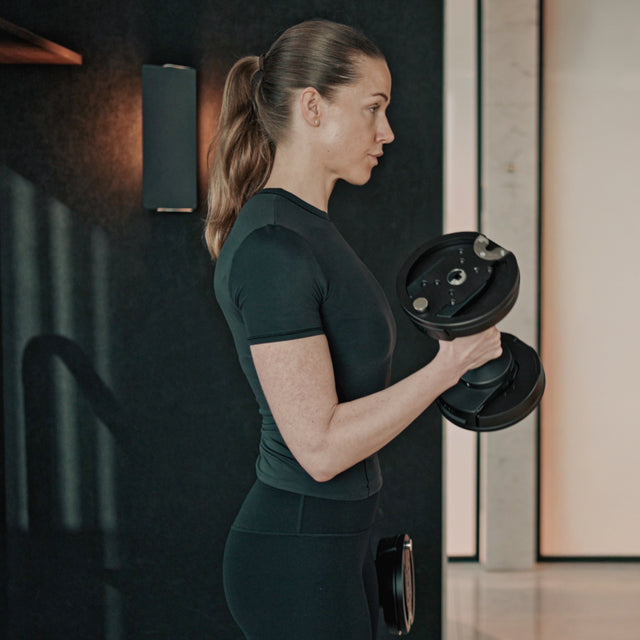A woman in a black workout outfit stands in profile, lifting a NÜOBELL-S Adjustable Dumbbell by Bells of Steel USA with one arm in a modern home gym.