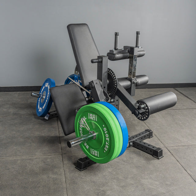 The Bells of Steel USA Legacy Leg Extension / Hamstring Curl Machine features a padded seat and rollers, plate-loaded with blue and green weights, and sits on a gray floor against a plain gray wall—ideal for home gyms.
