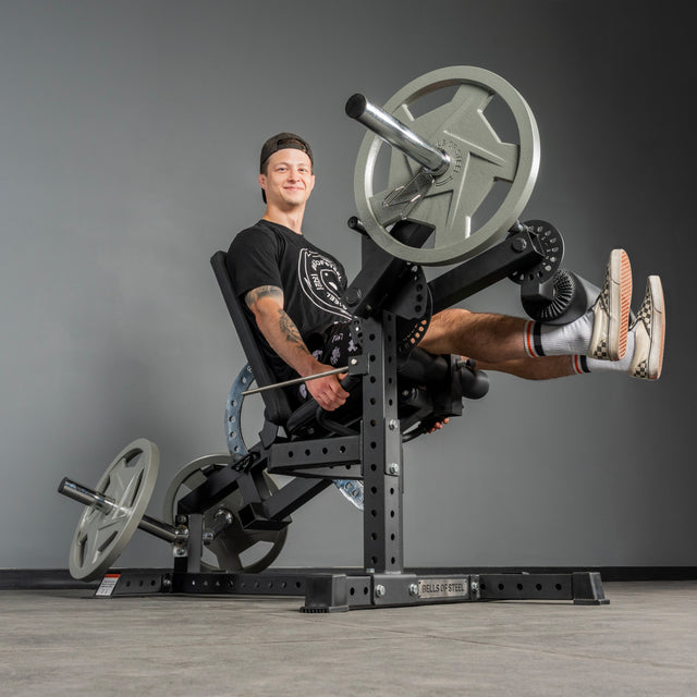 A man in a black t-shirt, shorts, and cap uses the Bells of Steel USA Legacy Leg Extension / Hamstring Curl Machine – Plate Loaded in the gym, smiling at the camera. This equipment is ideal for effective lower body workouts at home.