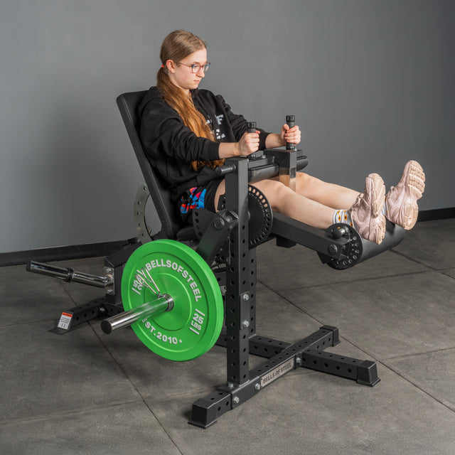 A woman in glasses and athletic wear uses the Bells of Steel USA Legacy Leg Extension / Hamstring Curl Machine – Plate Loaded, featuring a green weight plate, in a home gym with gray walls and flooring.