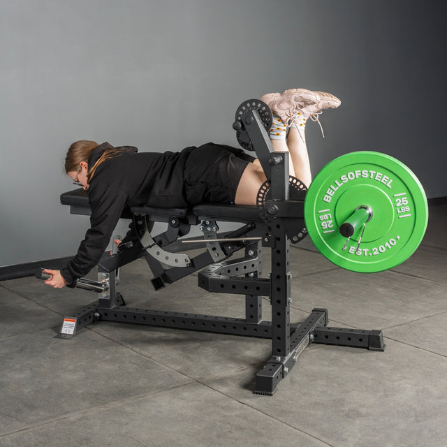 Wearing athletic gear, a person uses the Bells of Steel USA Legacy Leg Extension / Hamstring Curl Machine – Plate Loaded, performing a leg curl with a green 25 lb plate in an indoor gym with gray walls and tiled flooring.