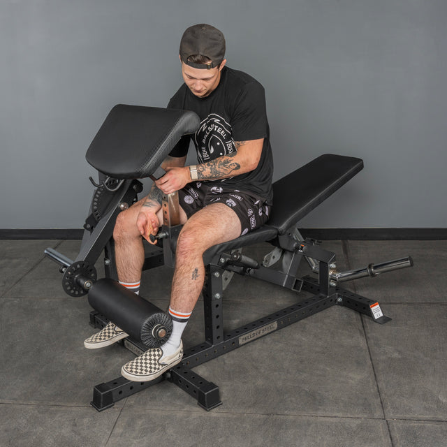A person in a cap, t-shirt, and shorts sits on a black adjustable bench, setting the Bells of Steel USA Legacy Leg Extension / Hamstring Curl Machine - Plate Loaded in a gym with gray walls and tiled floor.
