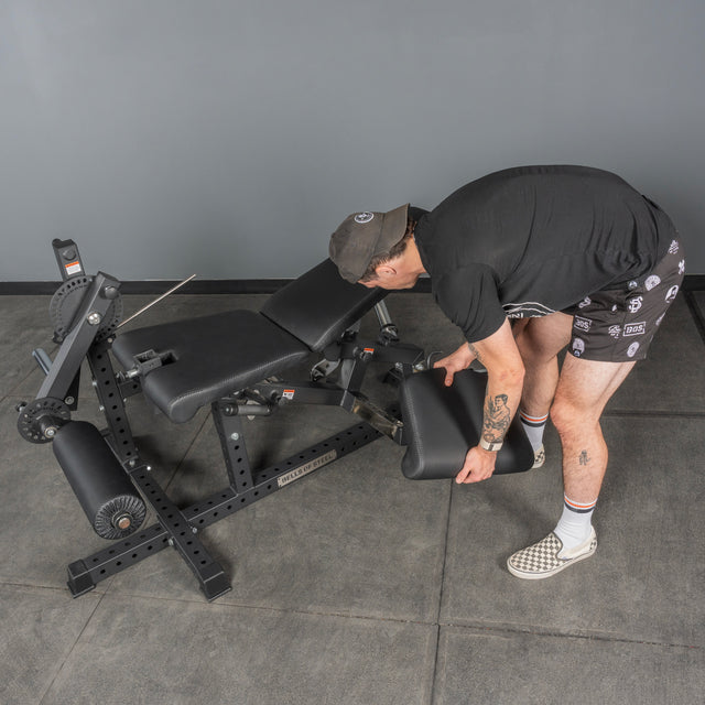 Wearing a cap, black t-shirt, patterned shorts, and checkered shoes, a person adjusts the footrest of a black workout bench in a home gym with gray walls—ideal to pair with the Bells of Steel USA Legacy Leg Extension / Hamstring Curl Machine.