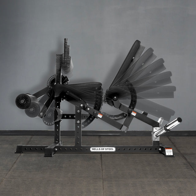 The Bells of Steel USA Legacy Leg Extension / Hamstring Curl Machine - Plate Loaded is shown on a gray floor and wall, with faded overlays highlighting the adjustable arm and pad positions for targeted exercises. Ideal for any home gym.