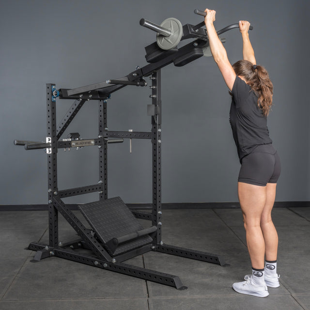 A woman in athletic wear stands before the Bells of Steel USA Pandemonium Squat – 3-in-1 Pendulum Squat / Calf Press / Viking Press Machine, reaching up to grip the handles as she prepares for an overhead exercise.