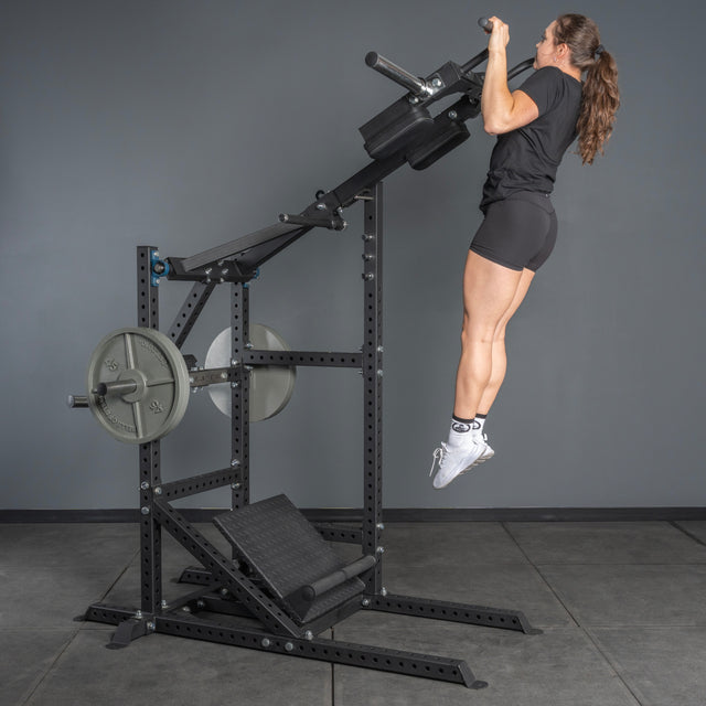 A woman in athletic wear uses a black pull-up bar machine with weights in a gym, next to the Bells of Steel USA Pandemonium Squat—3-in-1 Pendulum Squat / Calf Press / Viking Press Machine—for a multifunctional workout.