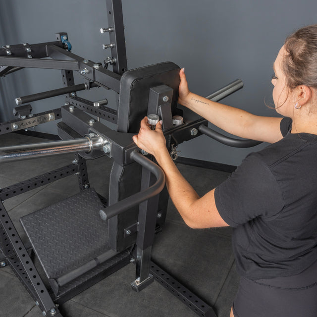A woman adjusts the seat of a Bells of Steel USA Pandemonium Squat – 3-in-1 Pendulum Squat / Calf Press / Viking Press Machine in a gym, standing on a black mat and wearing a black shirt. The machine features multiple handles and padded supports.