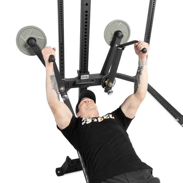 A person in a black shirt and cap uses the Bells of Steel Shoulder Boulder/Chest Fly Attachment, pressing weights upward on a chest press machine with metal plates beside a rack attachment against a plain white background.