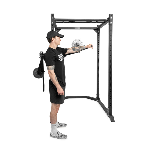 A man in black athletic wear stands sideways in a power rack, using the Bells of Steel Shoulder Boulder/Chest Fly Attachment for shoulder exercises, with one arm extended and a fan visible in the background.