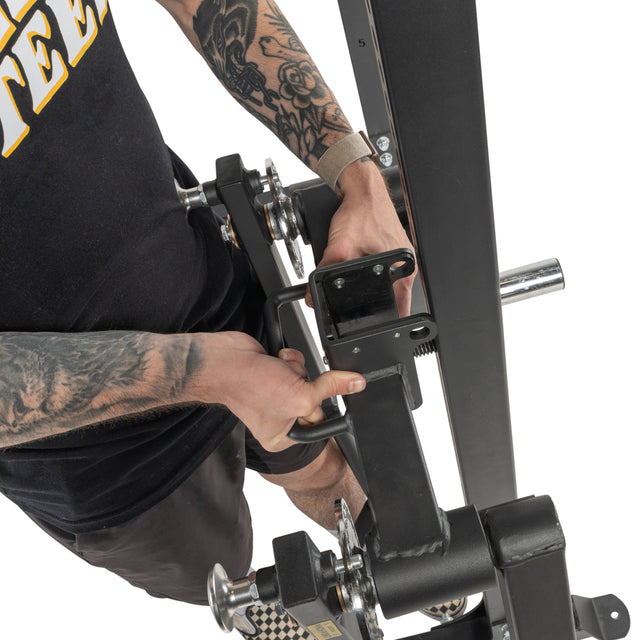 A tattooed person in a black shirt and shorts adjusts the handle of the Bells of Steel Shoulder Boulder/Chest Fly Attachment, viewed from above with metal and padded parts visible.