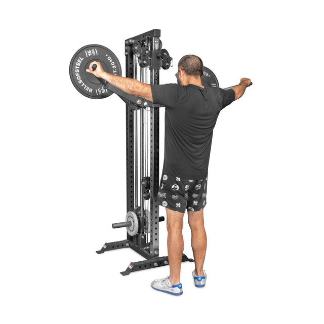 A man in black workout gear and blue-accented sneakers uses the Bells of Steel Shoulder Boulder/Chest Fly Attachment to perform a lateral raise. The background is plain white.