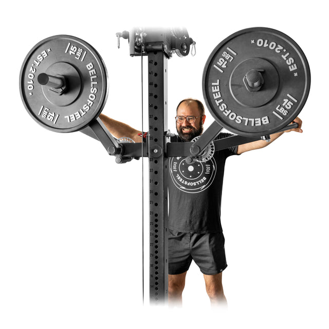A person wearing glasses and a black t-shirt stands behind the Bells of Steel Shoulder Boulder/Chest Fly Attachment, smiling with arms outstretched near two “Bells of Steel” weight plates—a perfect space-saving choice for your next shoulder workout.